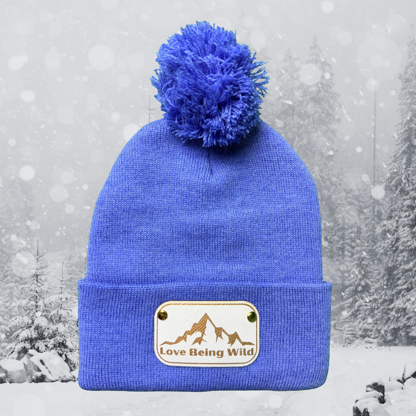 Blue beanie with pom-pom and 'Love Being Wild' patch against a snowy background