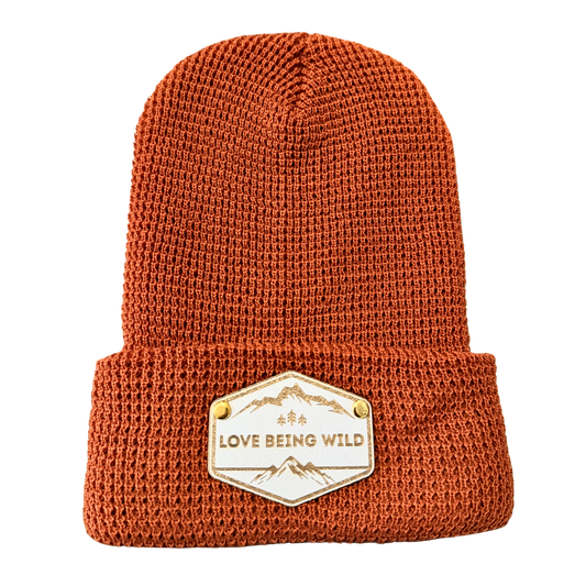 Brown knit beanie with 'Love Being Wild' patch on a white background