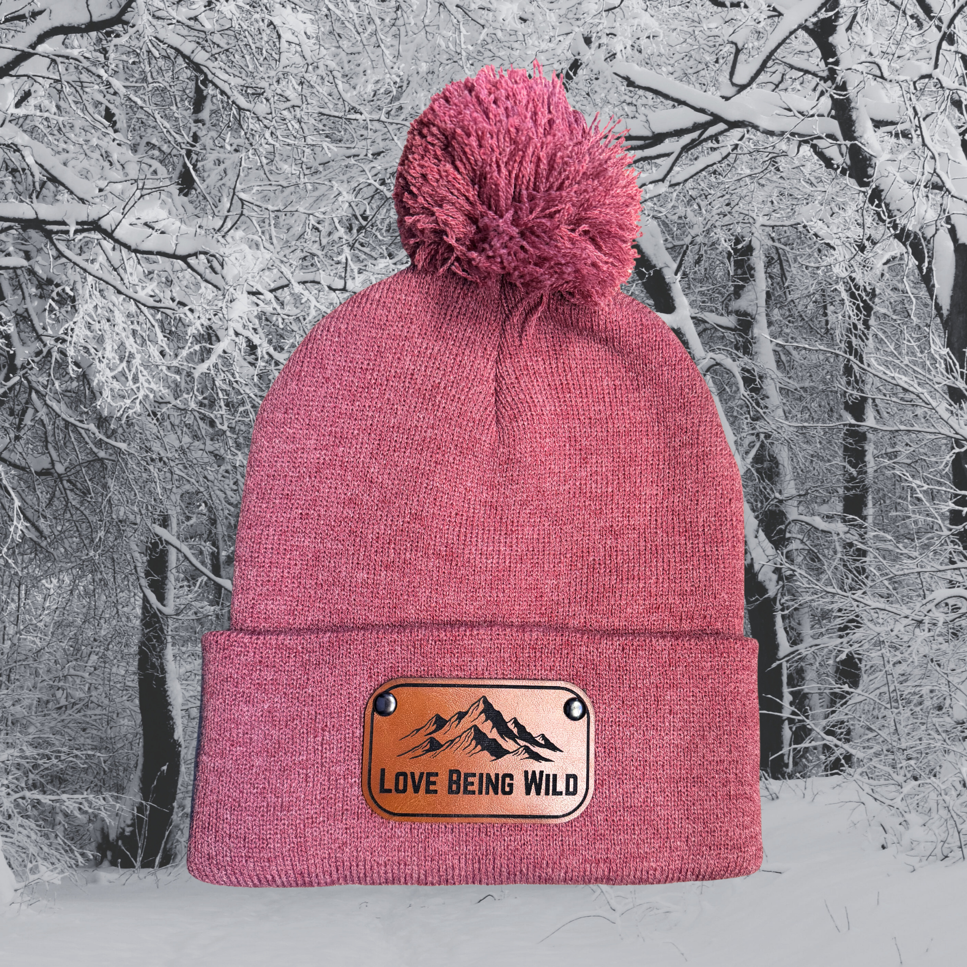 Pink beanie with a leather patch reading 'Love Being Wild' against a snowy forest background