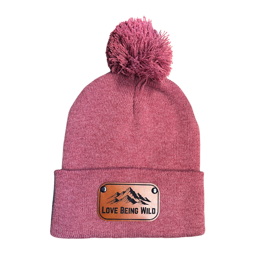 Pink beanie with a pom-pom and 'Love Being Wild' patch on a white background